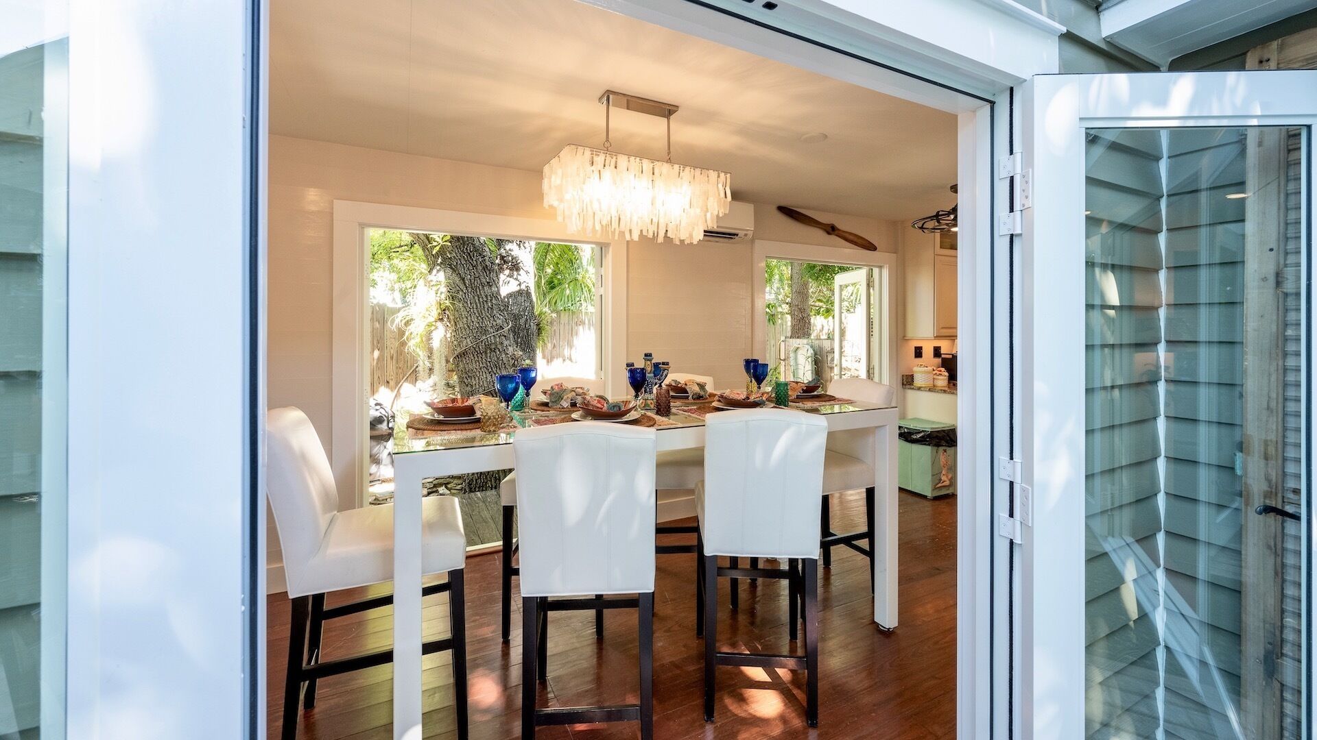 A dining area with a table set for four, surrounded by white chairs, a chandelier, and a view of greenery outside.