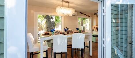 A dining area with a table set for four, surrounded by white chairs, a chandelier, and a view of greenery outside.