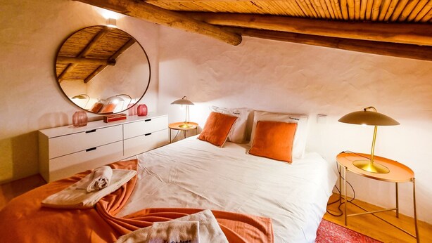 Een slaapkamer met een houten plafond, een ronde spiegel, een bed met wit en oranje beddengoed, een nachtkastje met lampen, en een dressoir.
