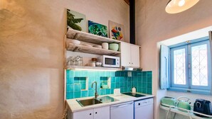 Een kleine keuken met een spoelbak, een magnetron en een vaatwasser. Boven de spoelbak zijn planken met servies en een magnetron. Een raam met tralies is rechts zichtbaar.