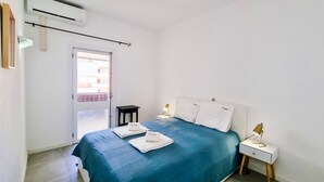 Une chambre à coucher avec un lit, des tables de chevet, un appareil de climatisation et une vue sur l’extérieur, visible à travers une porte.