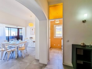 Una cocina moderna con armarios blancos, una zona de comedor con sillas blancas y una mesa preparada para cuatro personas, y vistas al exterior a través de una ventana.