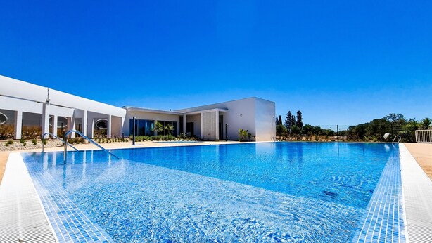 Um hotel moderno com uma grande piscina, passarelas cobertas e um céu azul límpido.