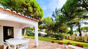 Eine Terrassenfläche mit weißem Tisch und Stühlen, eine überdachte Sitzgelegenheit und Blick auf einen gepflegten Garten mit Bäumen und Rasen.