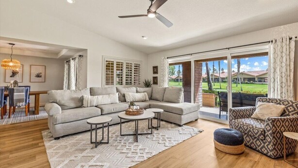 A spacious living room with a large sectional sofa, a patterned armchair, and a coffee table. There is a ceiling fan, a dining area with a table and chairs, and large windows offering a view of the outdoors. - Winter Escape in Chaparral Country Club (Palm Desert)