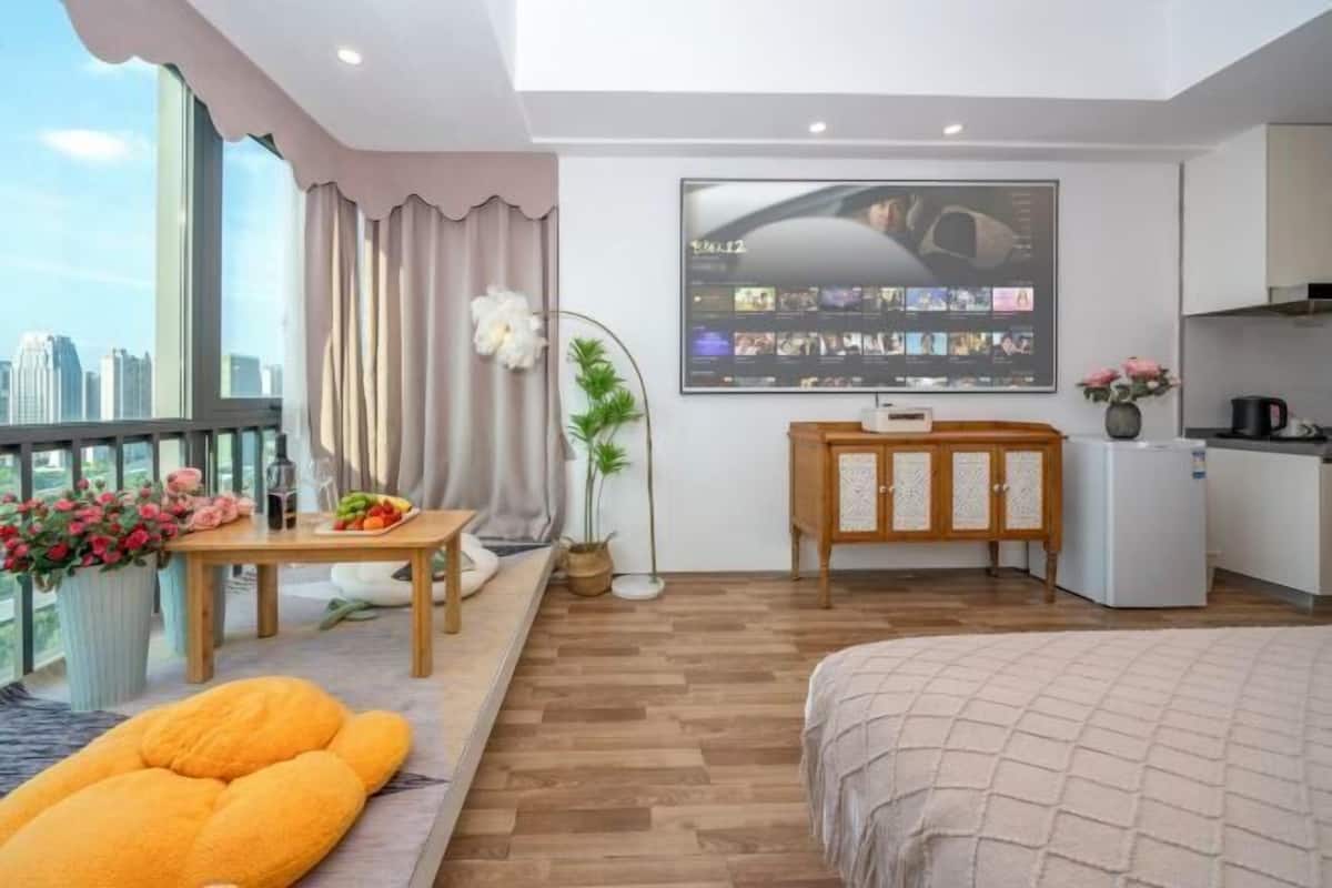 A modern hotel room with a large window, a flat-screen TV mounted on the wall, a wooden cabinet, and a bed with a beige blanket.