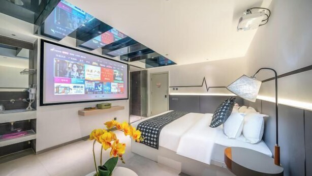 A modern hotel room with a large digital screen displaying content, a bed with white bedding, a bedside table with a lamp, and a vase with yellow flowers. - Zhiling Homestay (Five Star Street City Center) (Liuzhou)