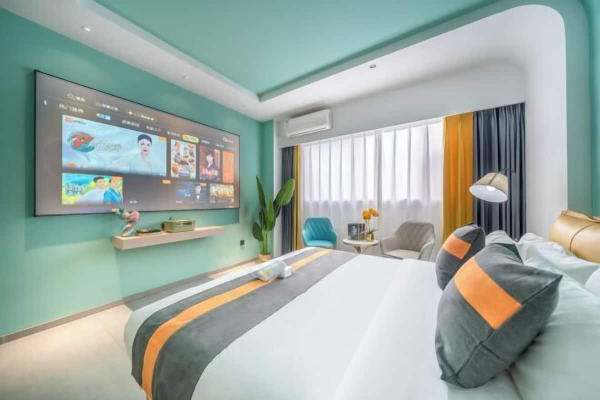 A modern hotel room with a large bed, a flat-screen TV mounted on the wall, a small side table, and a window with curtains.