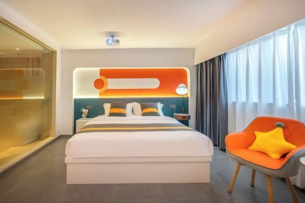 A modern hotel room with a large bed, a bedside table, a lamp, a chair, and a view of the outside through the window.