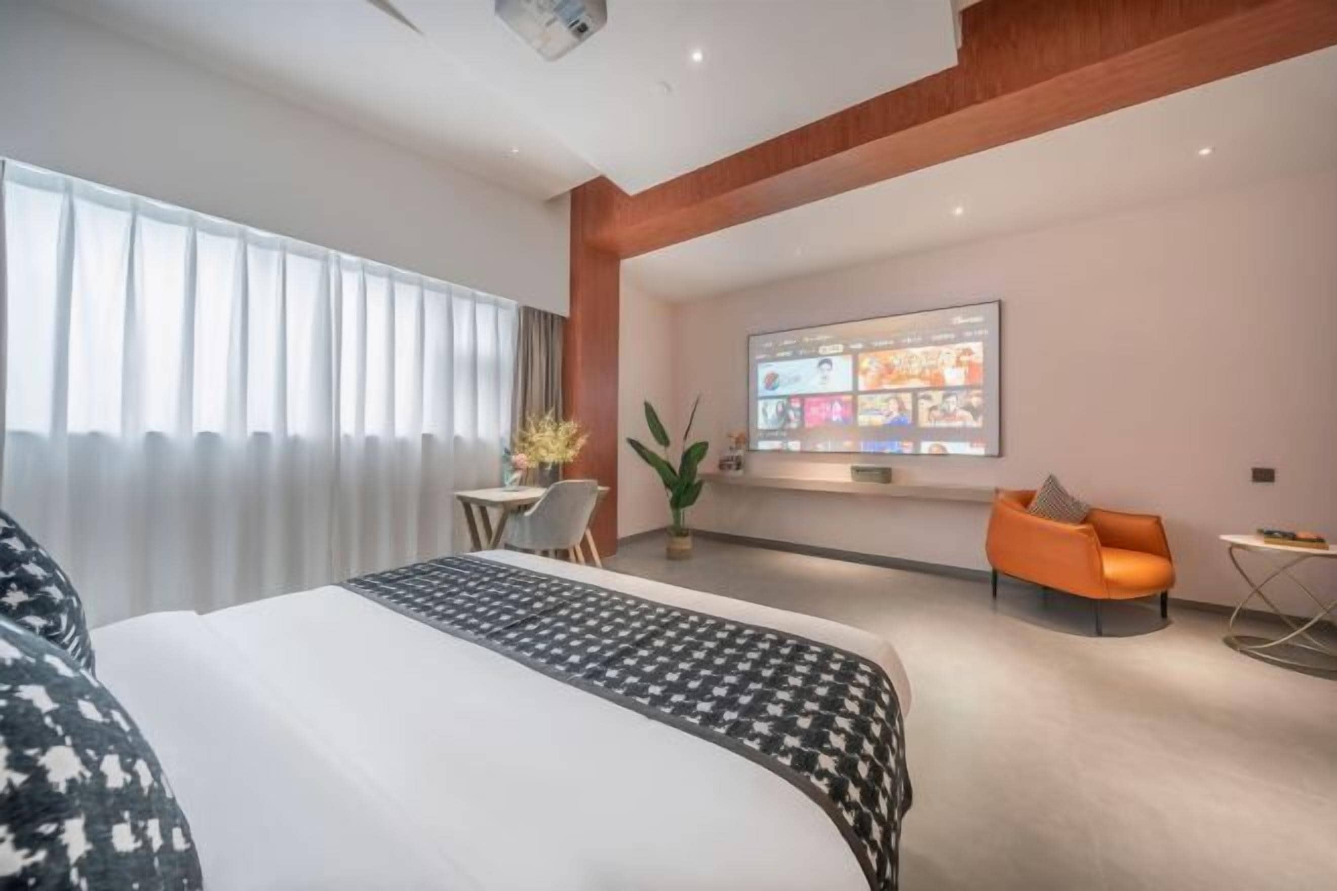 A modern hotel room with a large bed, a desk, an orange chair, and a TV displaying various channels.