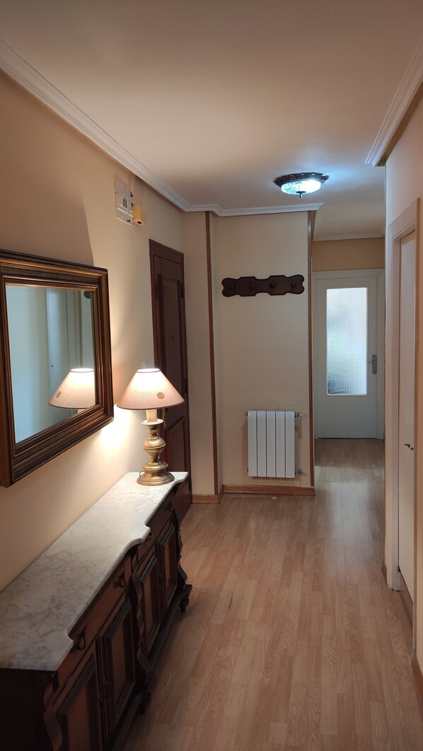A hallway with a wooden floor, a marble counter with a lamp, a mirror, a radiator, and a door leading to another room.