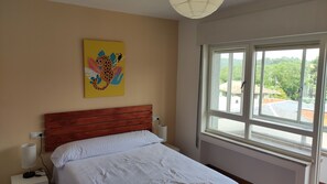 Une chambre à coucher avec une tête de lit en bois, un lit recouvert de linge de lit blanc, un tableau accroché au mur, une lampe de chevet et une fenêtre donnant sur un paysage verdoyant et des bâtiments.