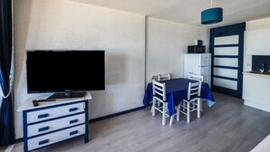 Un espacio habitable compacto con un televisor de pantalla plana sobre un mueble blanco, una mesa de comedor con sillas, y una zona de cocina con microondas y armarios.