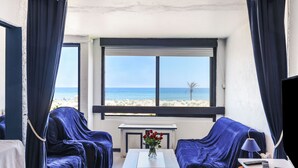 Una habitación con dos sillones tapizados en azul, una mesita con un jarrón de flores rojas, un televisor y una ventana que da a la playa y al océano.