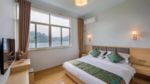 Ein Hotelzimmer mit großem Fenster, einem Bett mit vielen Kissen, einem Nachttisch und einer Wandklimaanlage.