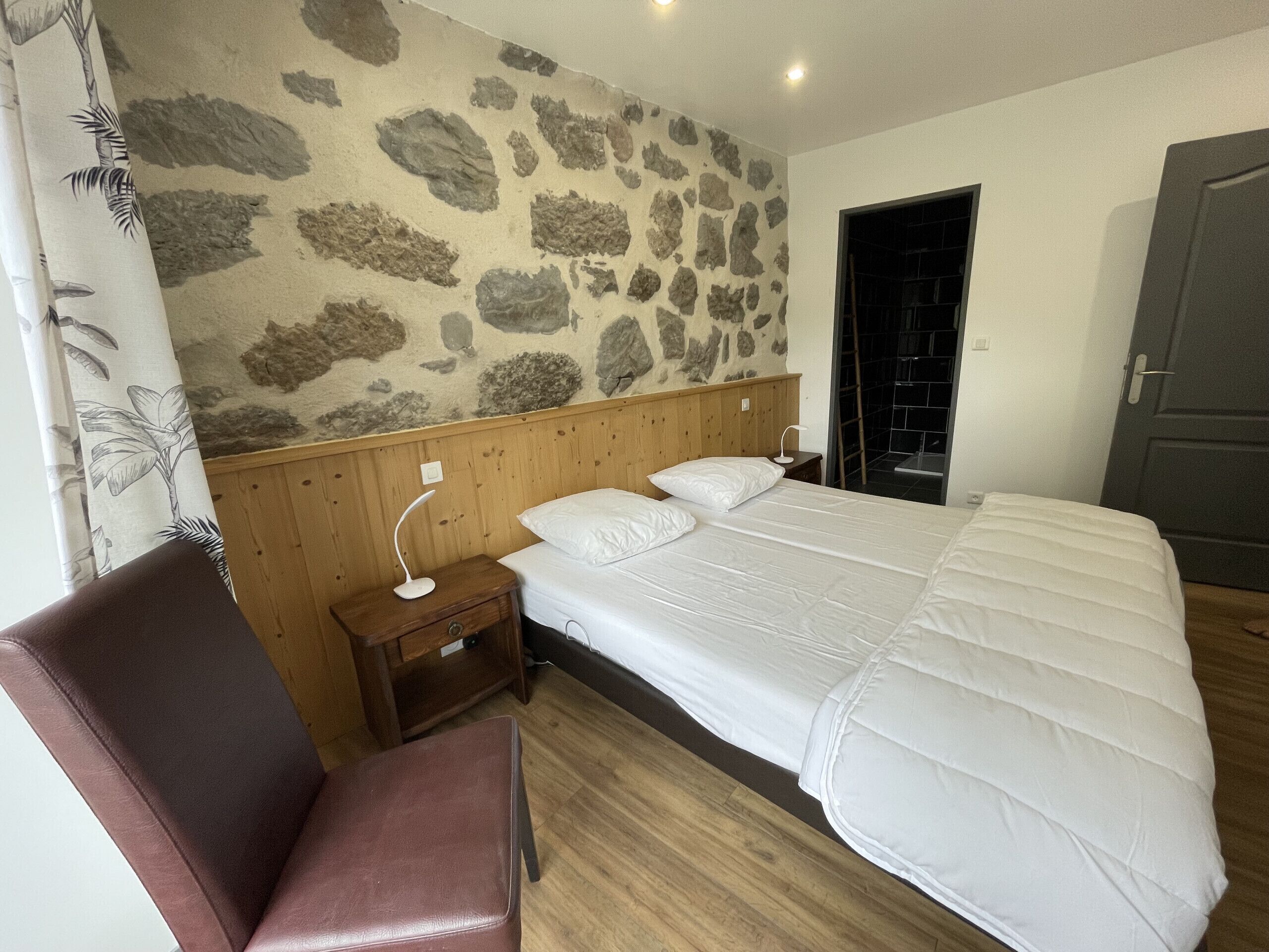 A bedroom with a stone-patterned wall, a wooden headboard, a bed with white bedding, a nightstand with a lamp, a brown leather chair, and a wooden floor.
