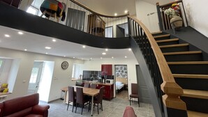Un vaste espace de vie comprenant une table à manger et des chaises, un escalier avec des rampes en bois, et une cuisine à l’arrière-plan.