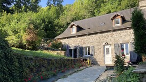 Une maison en pierre avec un chemin de pierre menant à une porte, entourée de verdure et d’un petit jardin agrémenté de fleurs rouges.