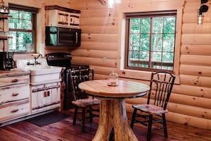 A rustic cabin interior with wooden walls, a round wooden table, two chairs, a kitchen area with a stove and microwave, and a window with a view of trees. - Cozy Treetop Getaway with Hot Tub (Millersburg)