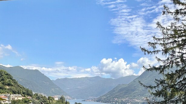 A balcony with a wooden bench overlooking a scenic view of a lake, mountains, and a town.