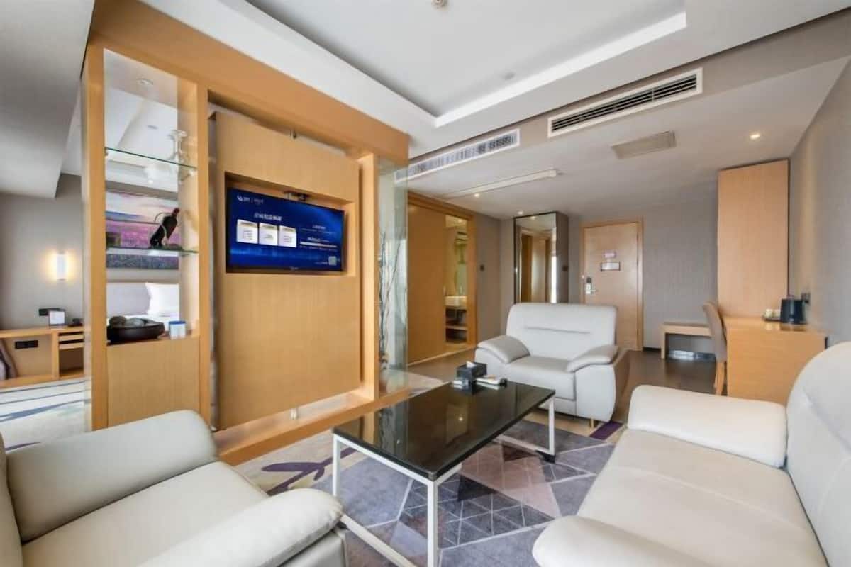 A modern hotel room with a glass partition, a flat-screen TV, a sofa, a coffee table, and a desk.