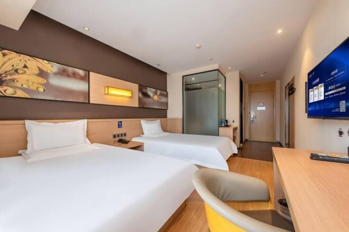 A hotel room with two beds, a desk, a chair, a TV, and a wall-mounted artwork.