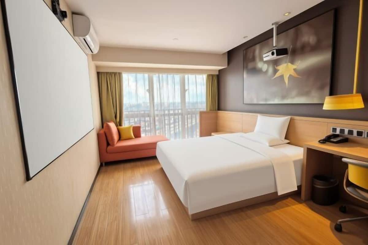 A hotel room with a bed, a desk, a chair, a sofa, a window with curtains, and a wall-mounted projector.