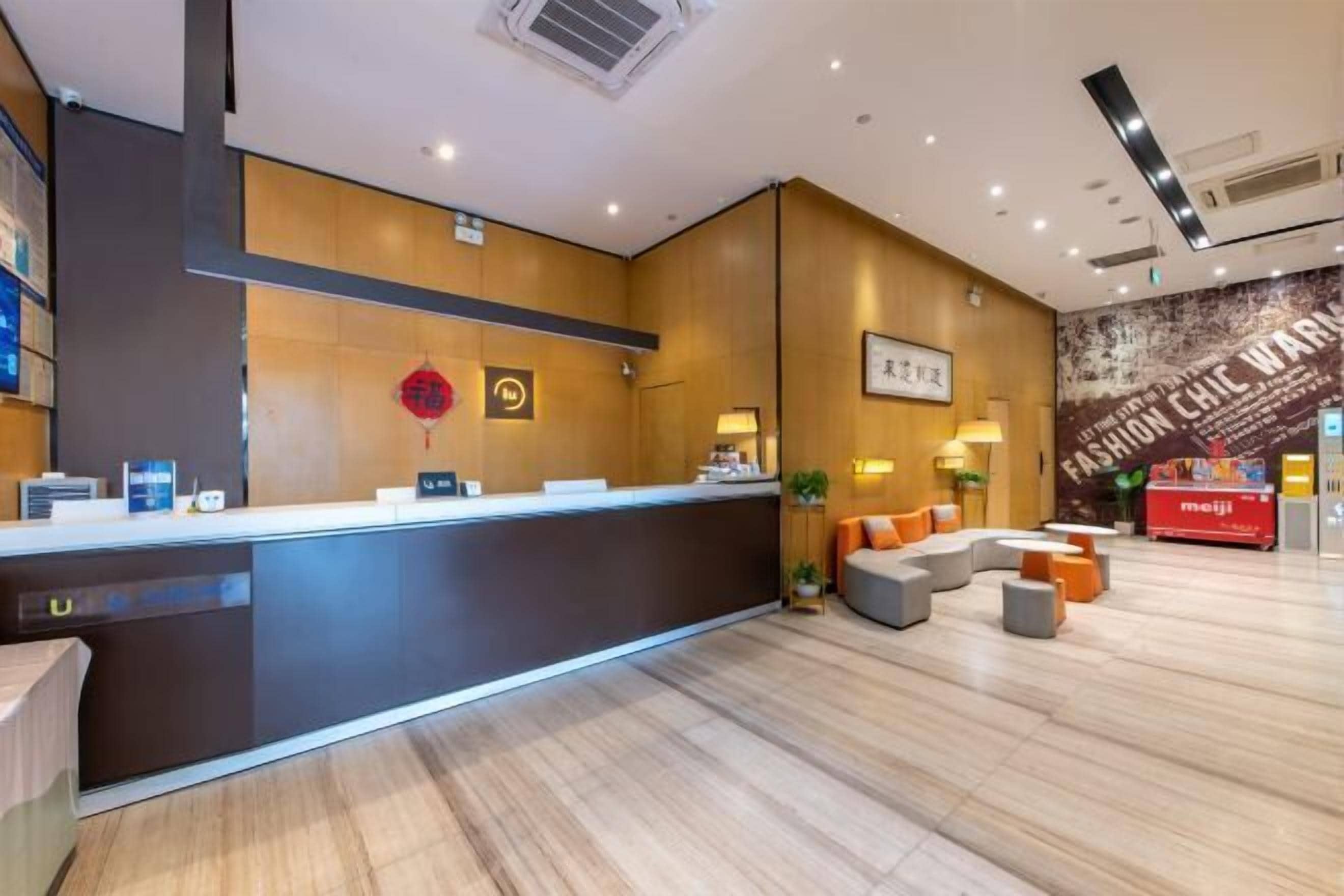 A modern hotel reception area with a wooden reception desk, a seating area with orange cushions, and a wall with a fashion-themed mural.