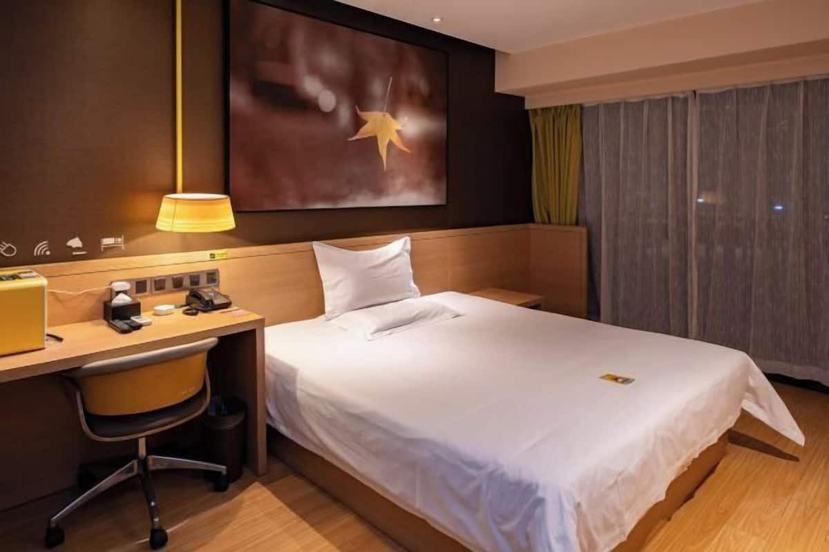 A hotel room with a bed, a desk with a lamp, a chair, a large artwork on the wall, and a window with curtains.