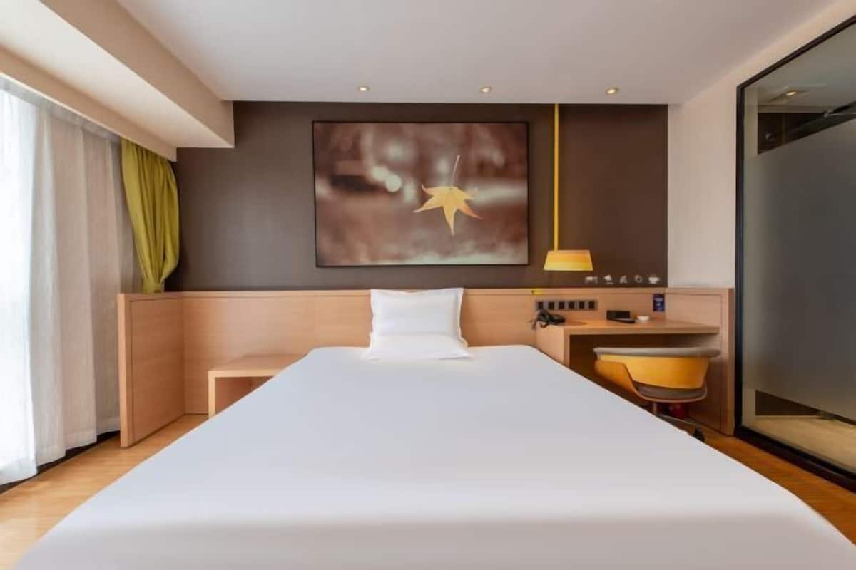 A modern hotel room with a large bed, a bedside table, a small desk, a chair, a lamp, and a large window with curtains.