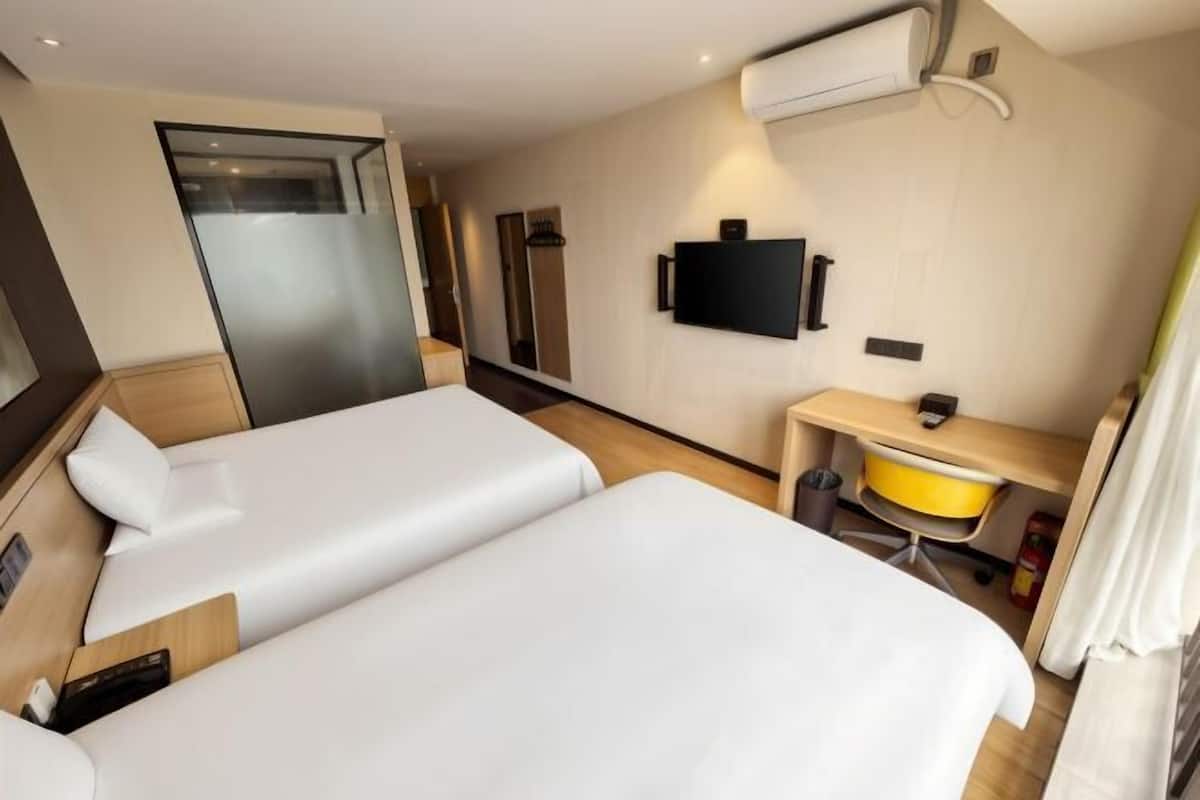 A hotel room with two beds, a desk, a chair, a TV, and an air conditioning unit.