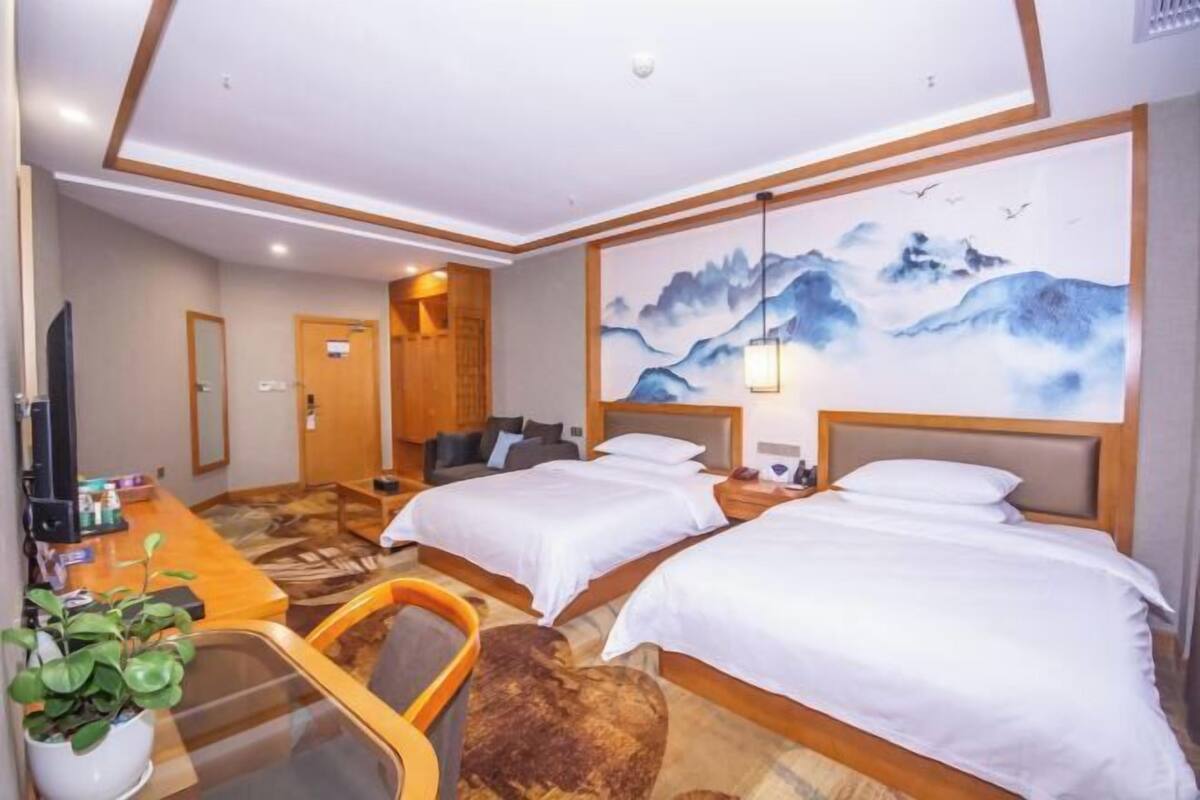 A hotel room with two beds, a desk with a chair, a TV, and a painting of mountains.