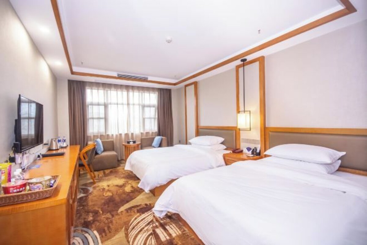 A hotel room with two beds, a desk with a TV, a sofa, and a large window with curtains.