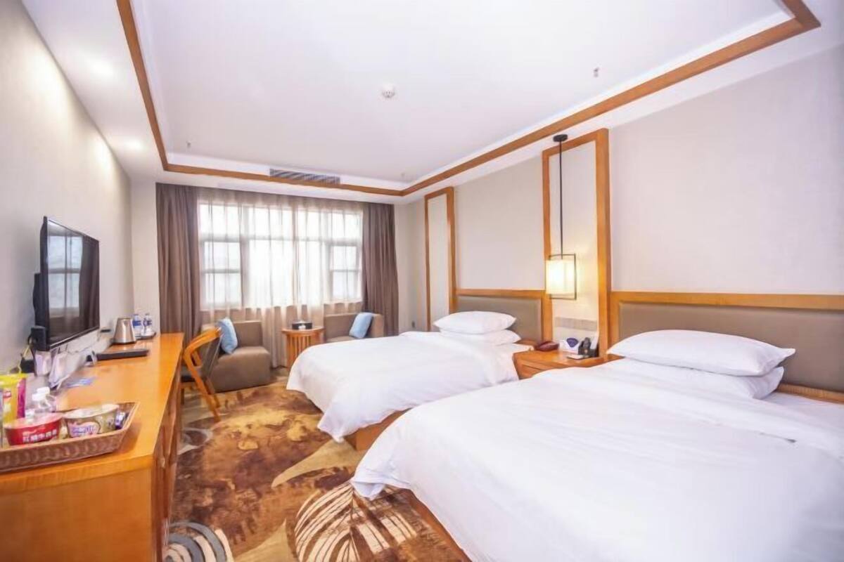 A hotel room with two beds, a desk with a TV, a sofa, and a large window with curtains.