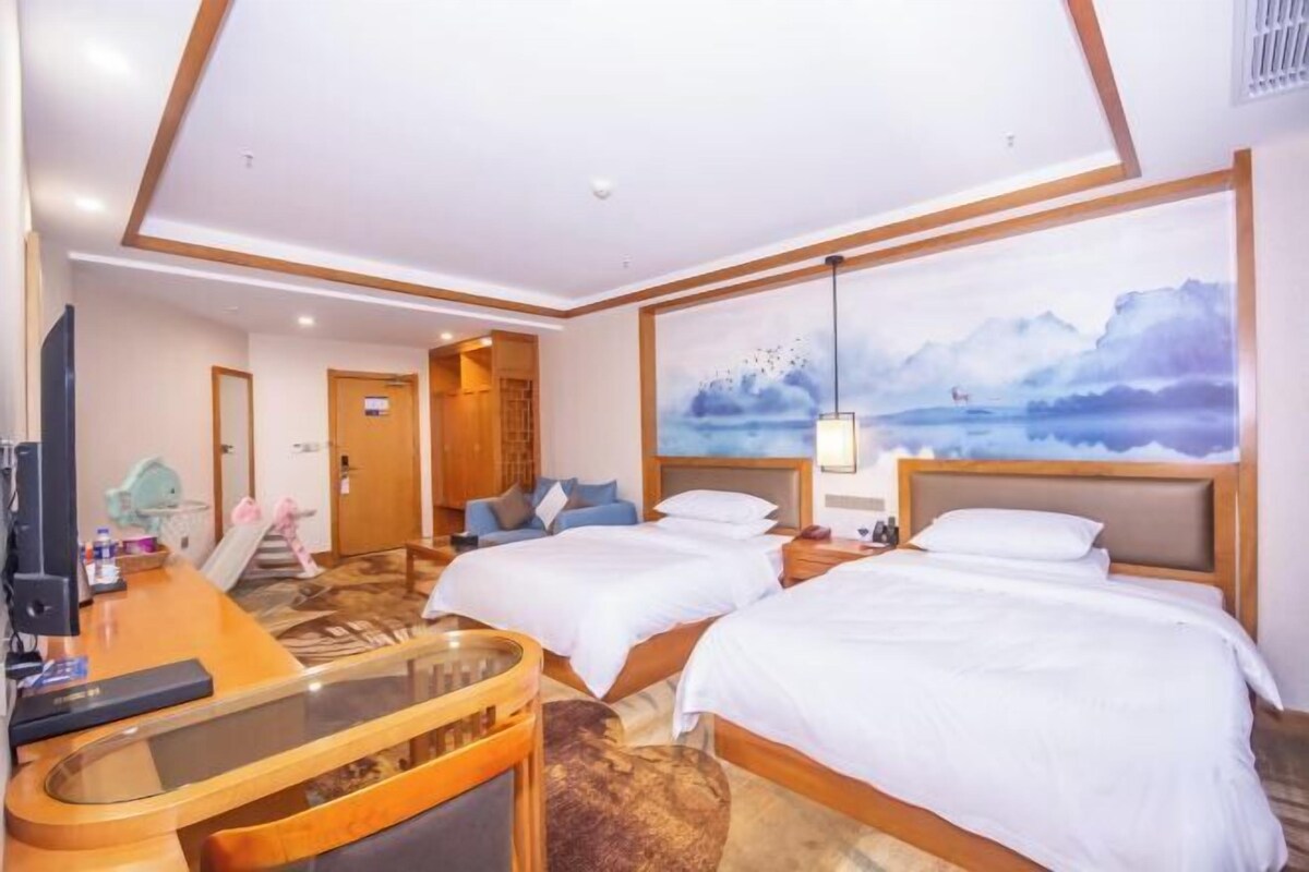 A hotel room with two beds, a desk with a glass top, a painting of a landscape, and a view of the sea.