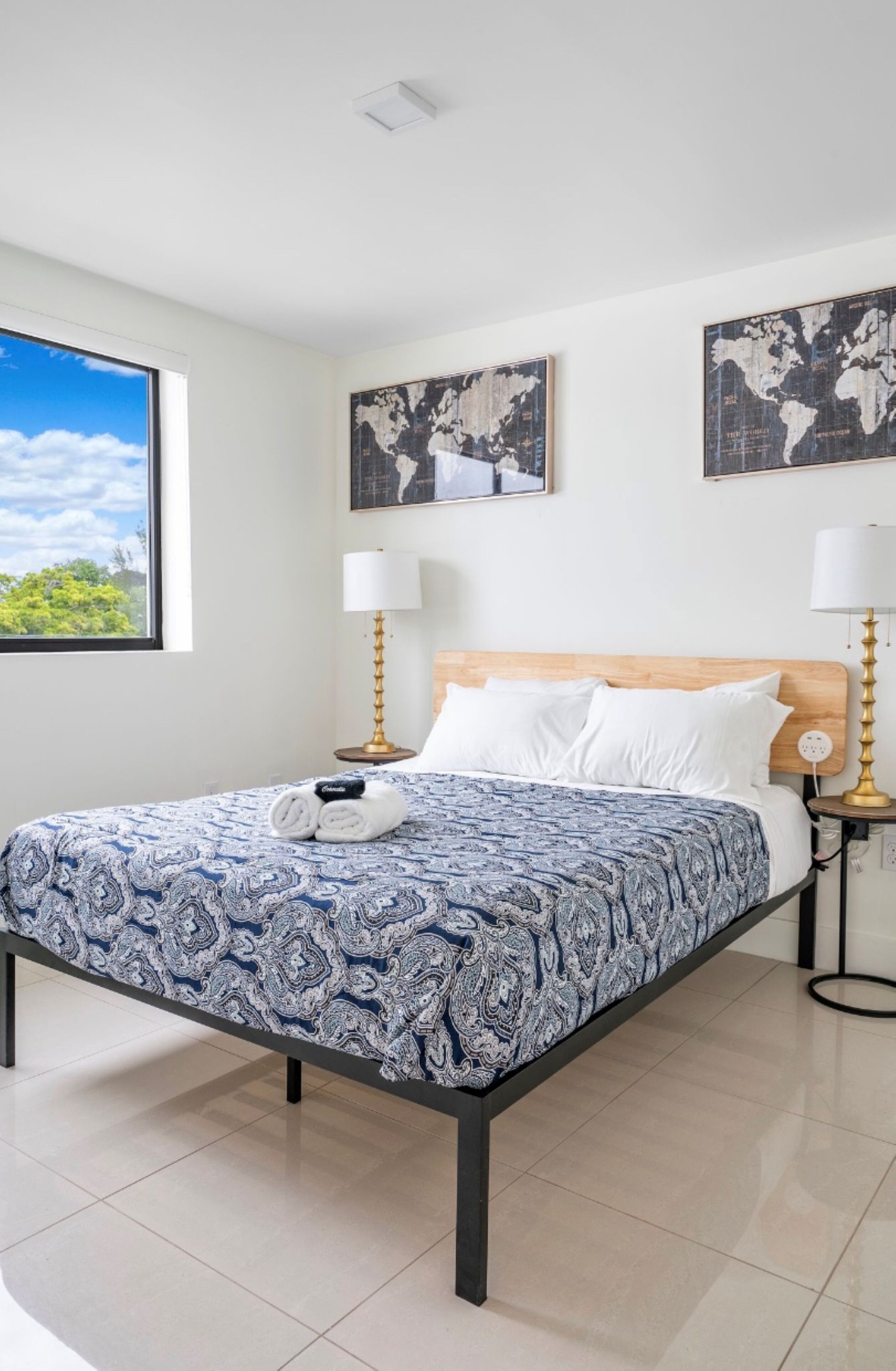 A neatly arranged bedroom with a large bed, two bedside lamps, a wooden headboard, and two world maps on the walls.