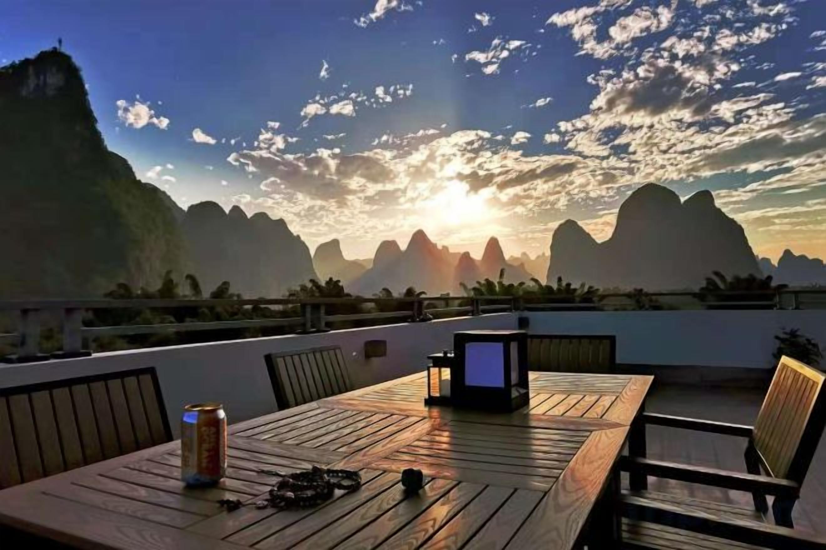 A wooden table with chairs, a laptop, and a can on a balcony overlooking a mountainous landscape.