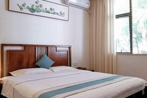 Ein ordentlich bezogenes Bett mit Holz-Kopfteil und blauem Kopfkissen, ein gerahmtes Blumenbild an der Wand und ein Fenster mit Vorhängen.