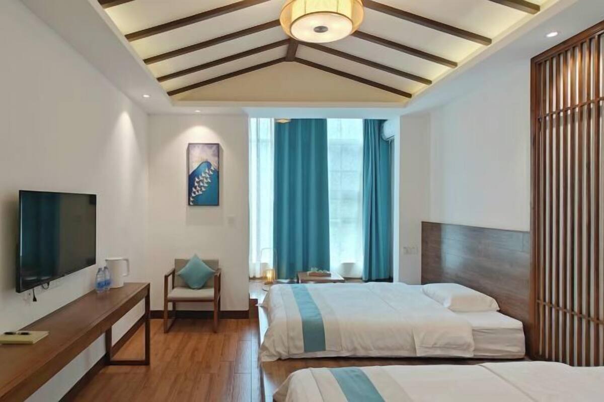 A modern hotel room with a wooden ceiling, a flat-screen TV, a bed with a striped bedspread, a chair, and a window with blue curtains.