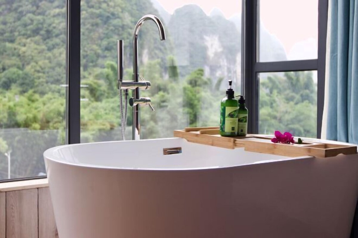 Ein modernes Badezimmer mit einer freistehenden Badewanne, einem Wasserhahn und einer Holzablage mit einer Flasche grüner Flüssigseife und einer rosa Blume.