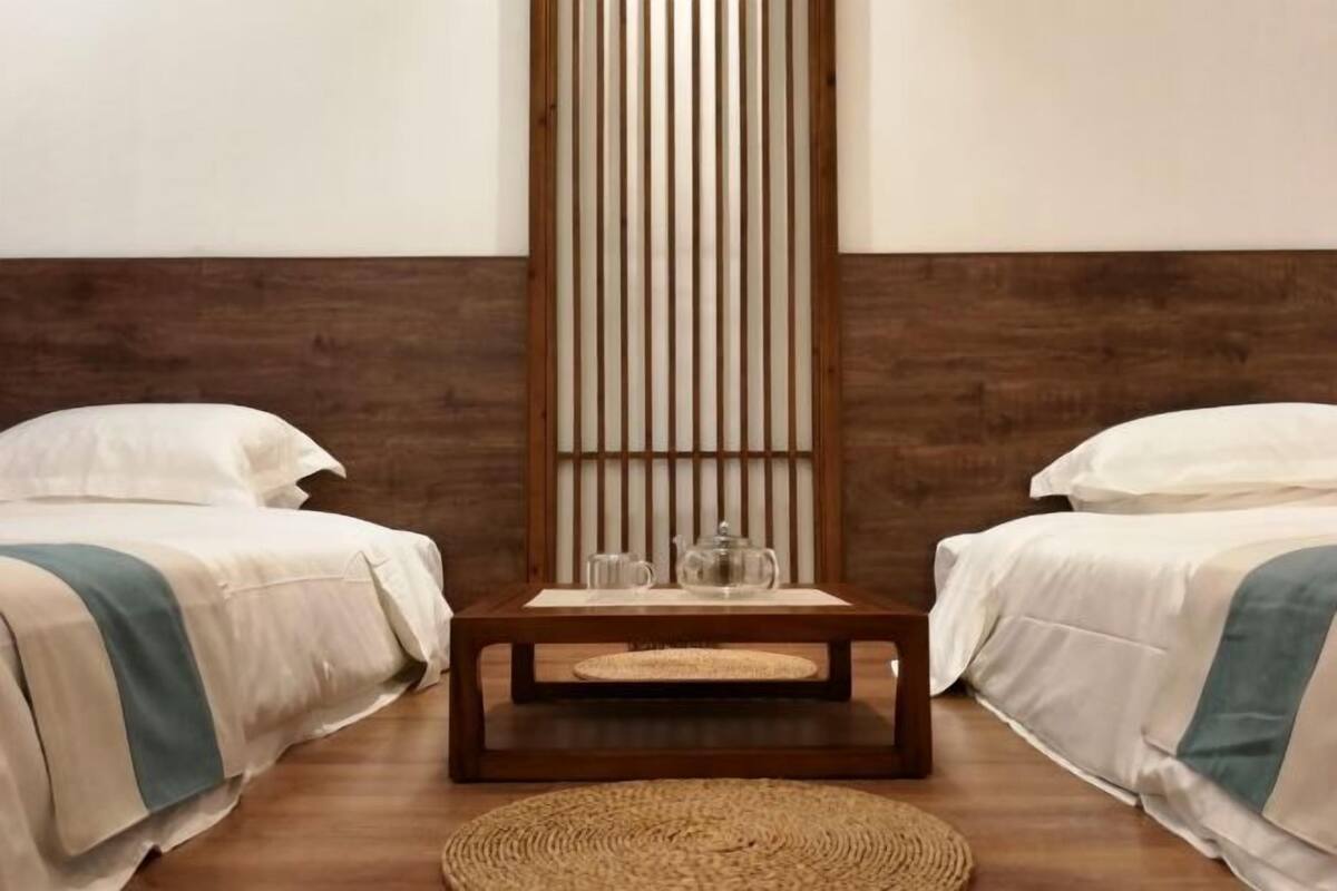 A room with two beds, a wooden floor, a woven rug, and a vertical wooden panel.