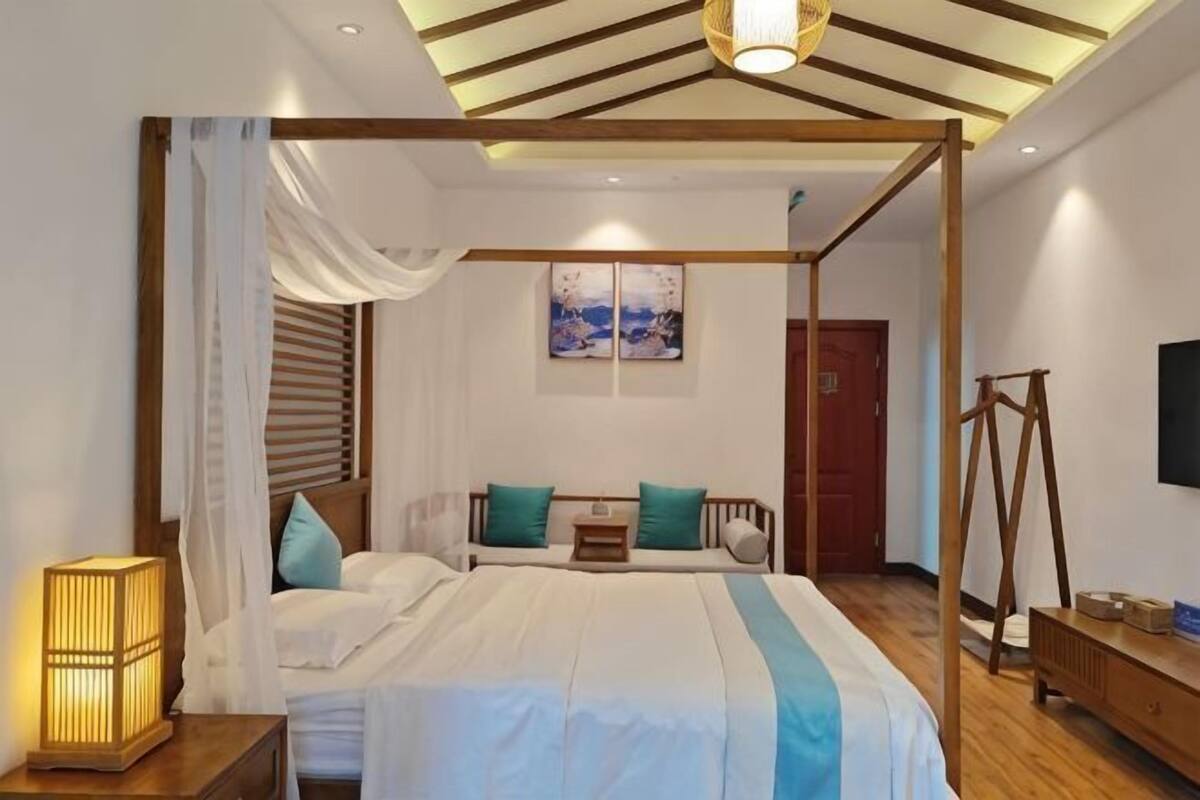 A four-poster bed with a canopy, a bedside table with a lamp, a wooden dresser, and a painting on the wall.
