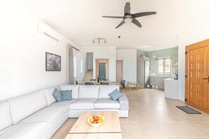 Un salon moderne avec un canapé blanc, une table basse en bois sur laquelle sont disposés des fruits, un ventilateur de plafond et une vue sur la cuisine.