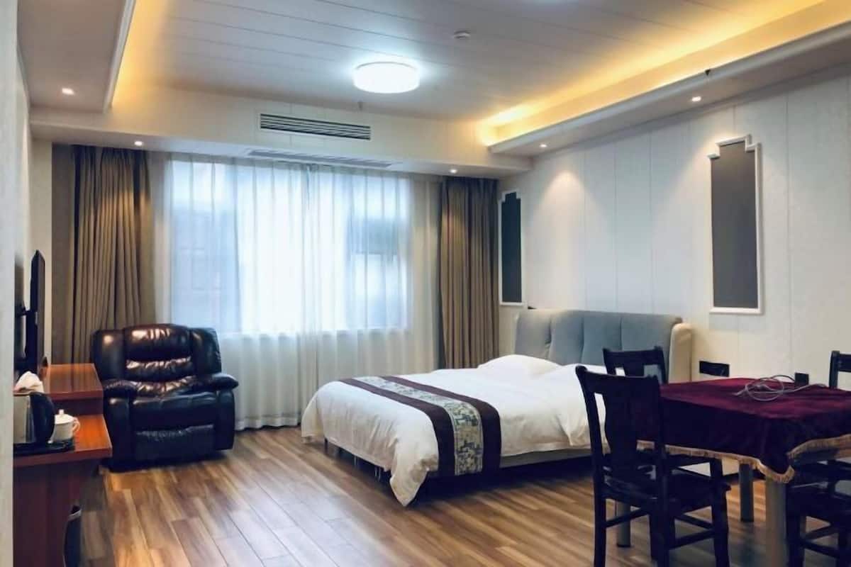 Quarto de hotel com cama, escrivaninha, poltrona de couro, mesa de jantar com cadeiras e uma grande janela com cortinas.