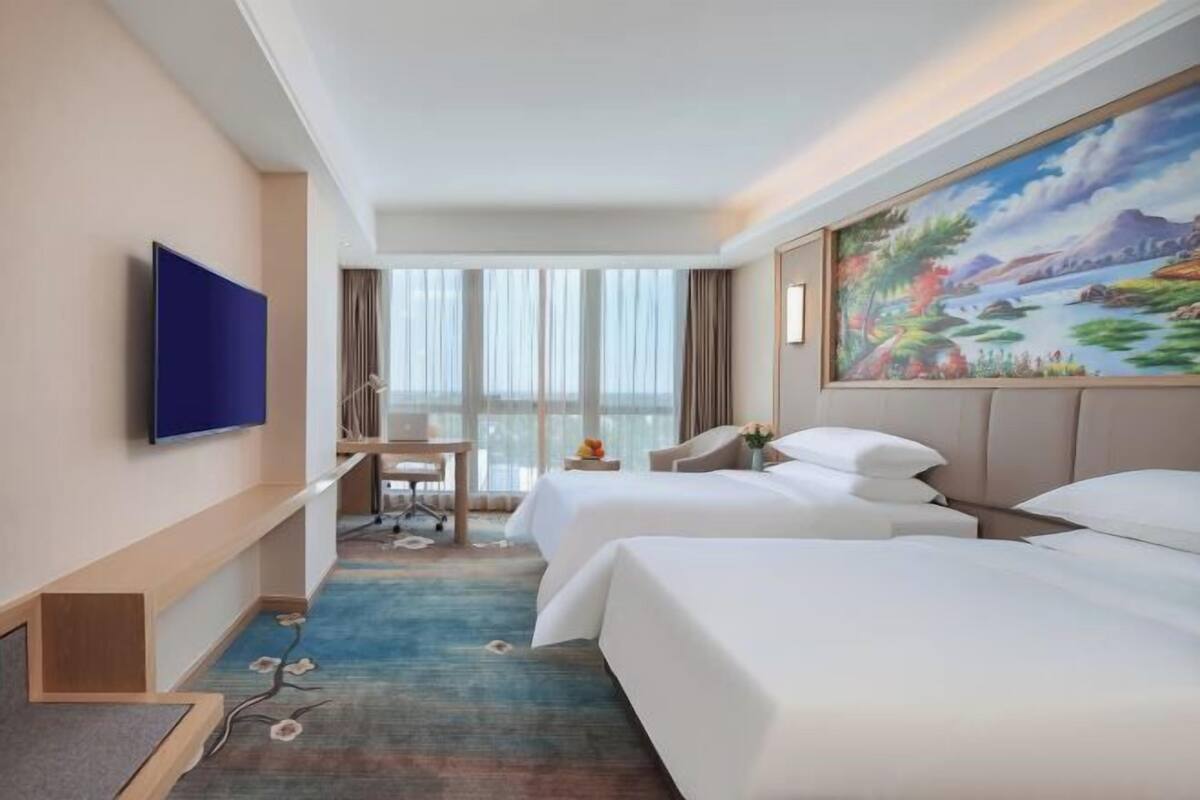 A hotel room with two beds, a desk, a TV, and a large window.
