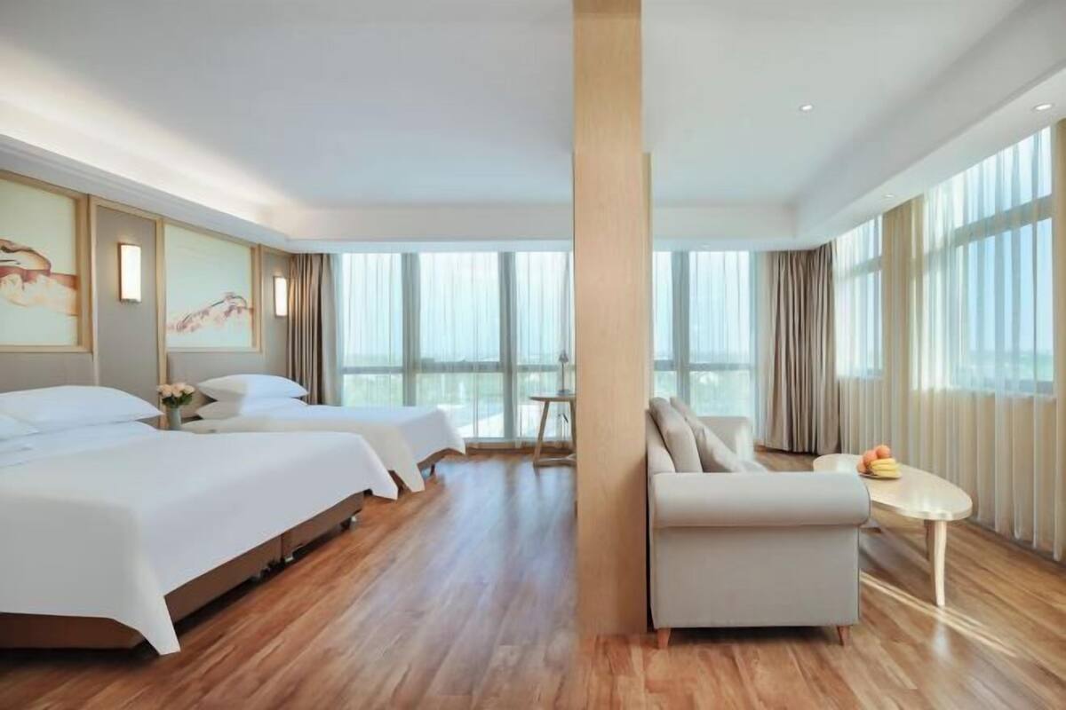 A hotel room with two beds, a sofa, a small table, and large windows.