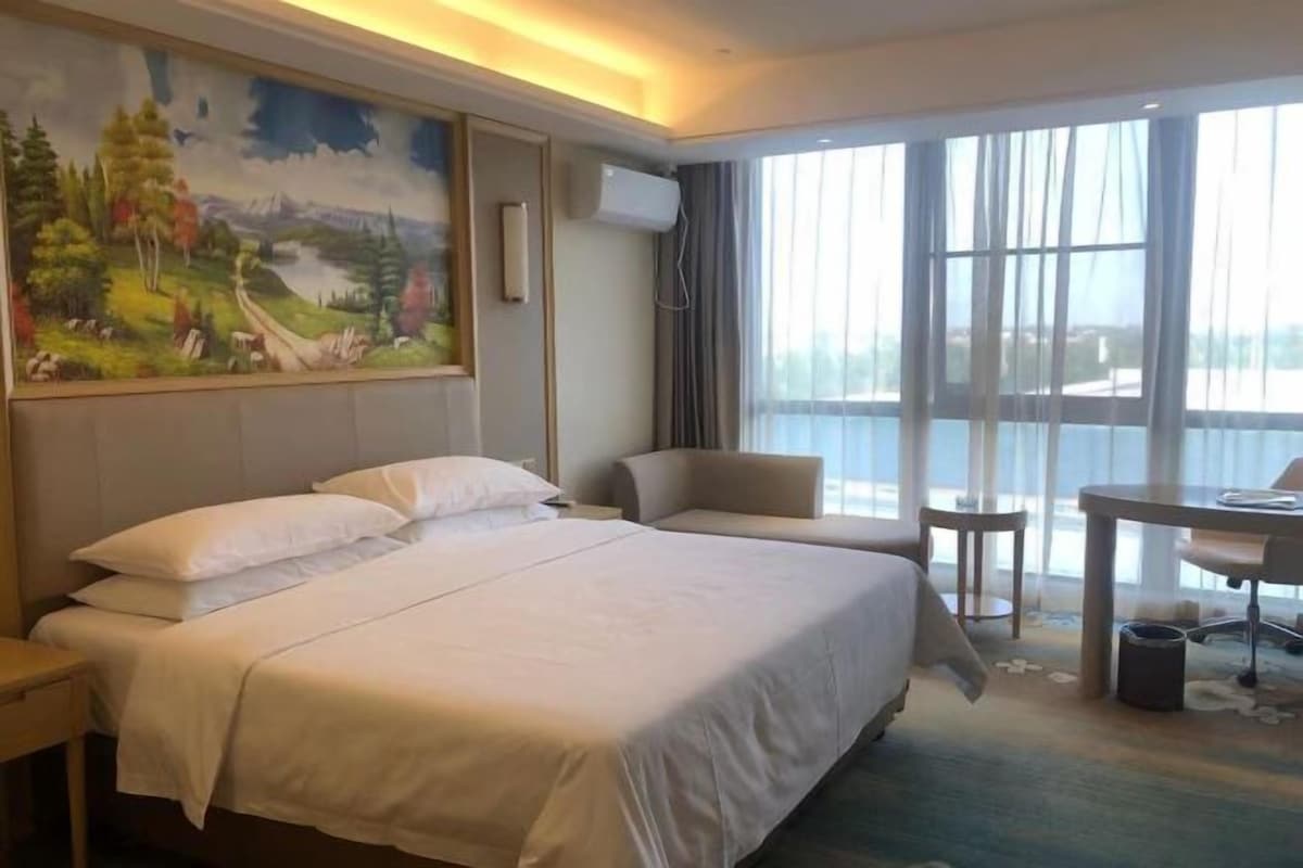 A hotel room with a large bed, a desk, a chair, and a view of the outdoors through large windows.