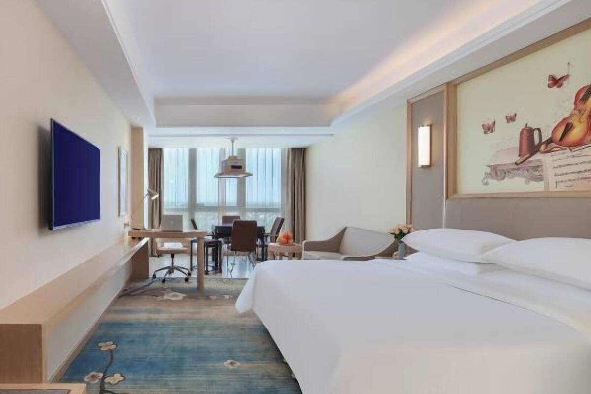 A hotel room with a large bed, a desk with a chair, a television mounted on the wall, and a window with curtains.