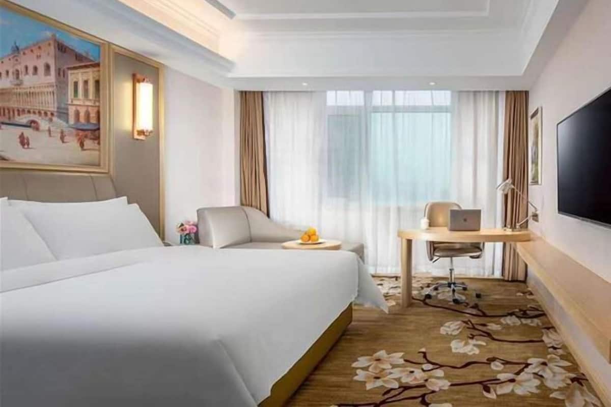 A hotel room with a bed, a desk with a laptop, a chair, a small table with fruit, and a painting of a cityscape.