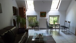 Ein Wohnzimmer mit einer Couch, einem Couchtisch und einem Essbereich mit Stühlen und einem Tisch. Es gibt Dachfenster, einen Deckenventilator und eine Heizung.